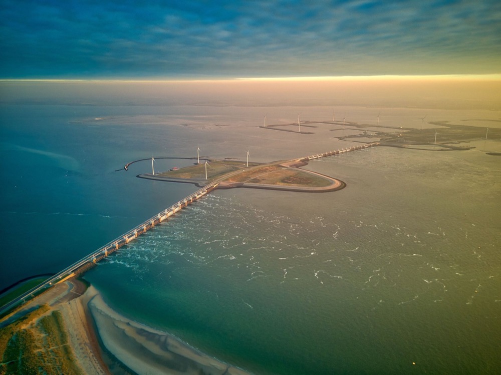 A long bridge and dam crossing the sea, with wind turbines along the shore, seen from the air