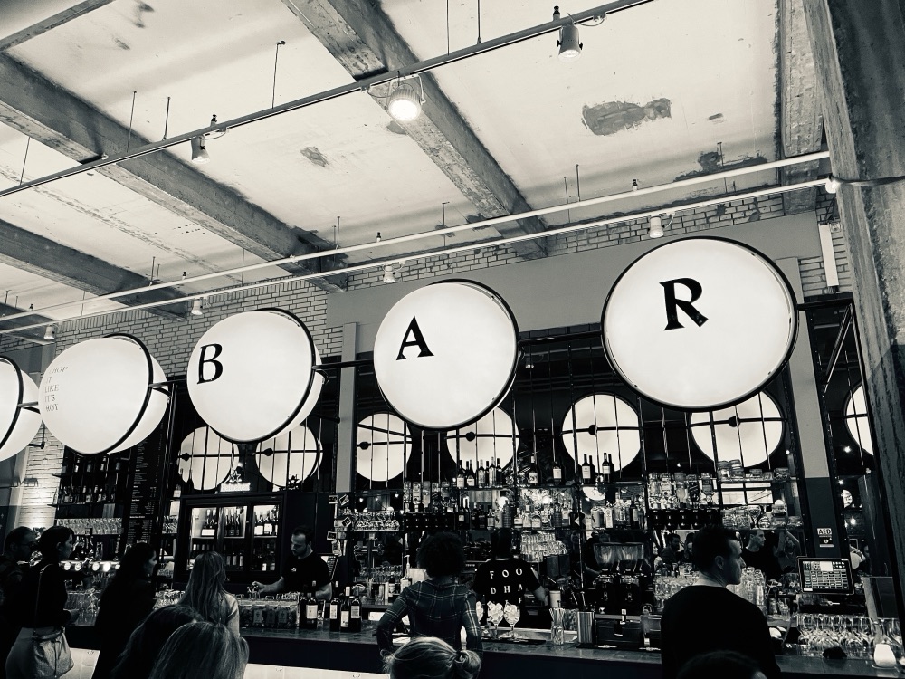 Large round illuminated letters spelling BAR above a market hall bar, in black and white